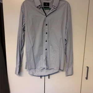 Ljusblå skjorta från Savvy Citizen - Klassisk ljusblå skjorta från Savvy Citizen med svarta knappar och diskret broderad SC-logga på ärmen. Skjortan har lång ärm, normal passform och är tillverkad i mjuk bomull. Perfekt för en clean och stilren look.