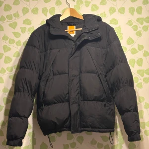 Svart pufferjacka från Timberland - Svart pufferjacka från Timberland med huva och dragkedja. Jackan har en boxig passform, stora vadderade paneler och dragsko nertill. Använd fåtal gånger så är som gott som ny:)