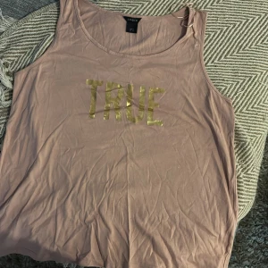 Beige linnetopp med guldtext från Lindex - Säljer en beige ärmlös linnetopp från Lindex i storlek M. Toppen har ett stort tryck med ordet 'TRUE' i glansig guldtext framtill. Mjuk och tunn bomullskänsla, perfekt för varma dagar eller lager på lager.