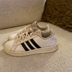 Adidas sneakers - Snygga vita sneakers från Adidas med klassiska svarta ränder på sidorna och logga baktill. Skorna har rund tå, platt sula och snörning framtill. Tillverkade i skinn med mjukt textilfoder för extra komfort. Perfekta för dig som gillar stilrena och sportiga skor.
