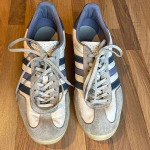 Adidas Gazelle sneakers blå/vit - Adidas Gazelle sneakers i vitt och blått med klassiska tre ränder på sidan. Ovandel i skinn och mocka, snörning framtill och gummisula med mönster. Snygg retrostil och diskret guldfärgad Gazelle-text på sidan. Perfekta för dig som gillar streetwear och klassisk adidas-look.