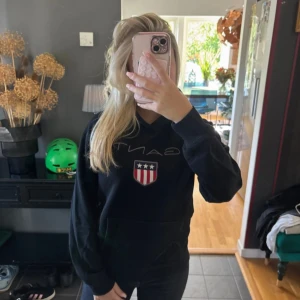 Svart hoodie från GANT med logga - Svart hoodie från GANT med stor broderad logga och sköld på bröstet. Klassisk känguruficka framtill och huva med snyggt randigt band på insidan. Perfekt för dig som gillar stilrena och sportiga plagg.