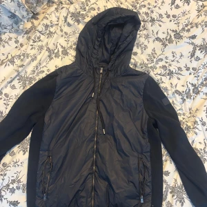 Svart vindjacka från BOSS med huva - Säljer en svart vindjacka från BOSS med huva och dragkedja framtill. Jackan har stickade ärmar och sidor, samt två fickor med dragkedja. Perfekt för dig som vill ha en stilren och sportig look. 