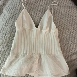 Vit ribbad linnetopp med knappar - Säljer en vit linnetopp med smala axelband och v-ringad front. Toppen har en ribbad mittdel och slät, lätt glansig nederdel med dekorativa knappar framtill. Materialmix av stickad och silkesliknande tyg ger en cool kontrast. Den har några sminkfläckar på insidan men inget som påverkar den när man har på sig toppen 🥰 