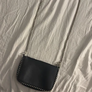 Svart axelväska från Stradivarius - Svart axelväska i skinnimitation från Stradivarius med silverfärgad kedjerem och nitar längs kanterna. Väskan har dragkedja upptill och en stilren, minimalistisk design som passar till många outfits.