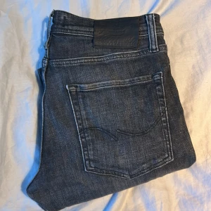 Svarta jeans från Jack & Jones - Snygga svarta jeans från Jack & Jones med klassisk femficksdesign och diskreta slitningar. Jeansen har raka ben och normal passform, tillverkade i mjukt denimtyg med tydliga sömmar och metallknappar. Perfekta för en stilren och avslappnad look.