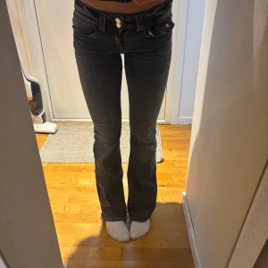 Svarta bootcut jeans med låg midja - Säljer ett par svarta jeans med bootcut-modell och låg midja. Jeansen har klassisk femficksdesign, dubbla knappar i midjan och är tillverkade i ett stretchigt jeansmaterial. Snygga att styla med crop top eller hoodie.