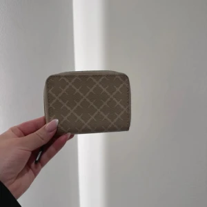 By Malena Birger Elia printed coin purse - Snygg beige plånbok med rutigt mönster och dragkedja runtom. Kompakt och smidig design som passar perfekt i väskan. Tillverkad i ett tåligt material med stilrena detaljer.