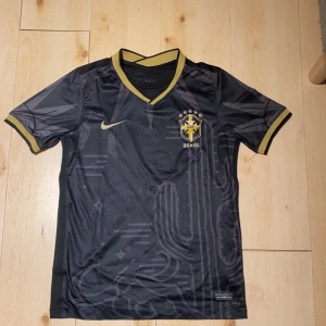 Svart Brasilien fotbollströja Nike - Snygg svart Brasilien fotbollströja från Nike med guldiga detaljer och CBF-emblem på bröstet. Tröjan har korta ärmar, V-ringad krage med gul kant och diskreta grafiska mönster på framsidan. Tillverkad i lätt och ventilerande material, perfekt för match eller träning.