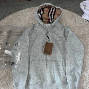 Grå hoodie från Burberry med dragkedja - Snygg ljusgrå hoodie från Burberry med klassiskt rutigt foder i huvan. Tröjan har dragkedja framtill, snörning i huvan och två stora fickor. Perfekt för dig som vill ha en stilren och lyxig look med streetkänsla.