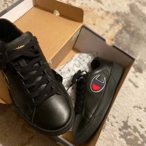 Svarta sneakers från Champion - Svarta sneakers från Champion med platt sula och rund tå. Skorna har snörning och är tillverkade i skinnimitation. På sidan finns ett stort Champion-logo i rött, vitt och blått. Snygg och stilren design som passar perfekt till streetwear. Skorna är helt nya, inte använda en endaste gång! Lådan och prislappen som fortfarande sitter på! 899kr nypris, jag säljer skorna för 400kr. Skorna är till män men kan absolut också passa kvinnor. Storlek 41! Priset kan diskuteras! Endast seriösa köpare, tack! 
