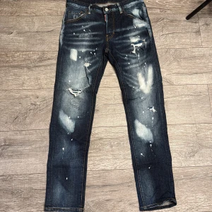 Mörkblå Dsquared2 jeans med stänk - Säljer ett par mörkblå jeans från Dsquared2 med coolt stänkmönster och slitningar på benen. Jeansen har raka ben, klassiska fickor och detaljerad söm. Bak finns en stor Ciao Bella-patch och tryck ovanför ena bakfickan. Perfekt för dig som gillar streetstyle.