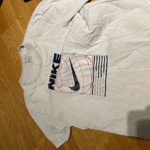 Vit Nike Dri-Fit t-shirt med tryck - Vit t-shirt från Nike med stort grafiskt tryck framtill i svart och rött. Klassisk rund halsringning och korta ärmar. Tillverkad i Dri-Fit material som andas och passar perfekt för träning eller chill. Snygg och sportig vibe.