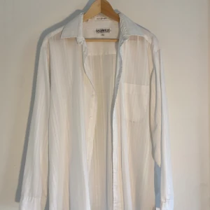 Vit randig skjorta från Hagenfeldt - Snygg vit skjorta från Hagenfeldt Collections i storlek L. Skjortan har diskreta vertikala ränder, klassisk krage och en bröstficka. Tillverkad i en mjuk mix av bomull och polyester, med knappar framtill och långa ärmar. Perfekt för dig som gillar stilrena och fräscha plagg.