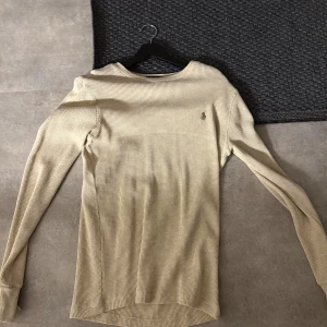 Beige långärmad tröja från Polo Ralph Lauren - Snygg beige långärmad tröja från Polo Ralph Lauren med diskret broderad logga på bröstet. Tröjan har ribbade muddar och är tillverkad i mjuk bomull med ett tunt, ribbat mönster. Säljs pga att den aldrig används och är småt för stor