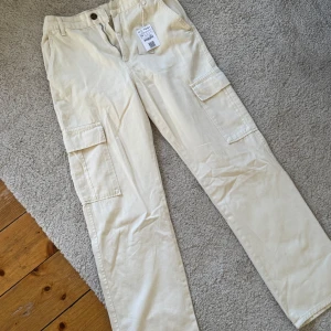 Beiga cargopants i bomull från Kappahl - Stilrena beiga cargopants från Kappahl i 100% bomull. Byxorna har raka ben, flera praktiska fickor på sidorna och bak, samt dragkedja och knapp framtill 