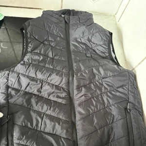 Svart värmeväst med USB och dragkedja - Svart dunväst med värmefunktion, USB-anslutning och powerknapp på bröstet. Quiltad design med snedställda sömmar, hög krage och hel dragkedja framtill. Perfekt för kalla dagar när du vill hålla dig varm och ändå se chill ut.