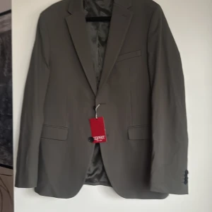 Grå kavaj slim fit från Esprit - Snygg grå kavaj från Esprit i slim fit-modell. Klassisk design med två fickor fram, bröstficka och knappar vid ärmslut. Tillverkad i polyesterblandning med slät finish och helfodrad insida. Perfekt för dig som vill ha en stilren och modern look.