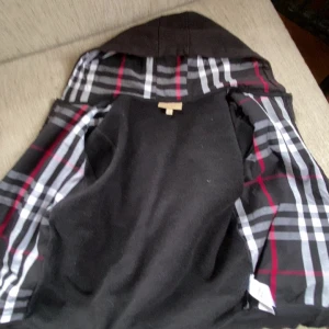 Svart Burberry hoodiejacka med rutig huva - Svart hoodiejacka från Burberry med dragkedja, broderad logga på bröstet och klassiskt rutigt foder i huvan. Jackan har två fickor framtill och är tillverkad i mjuk bomull. Perfekt för dig som vill ha en stilren och ikonisk look.