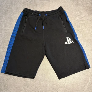 Svarta PlayStation shorts från H&M - Svarta shorts från H&M med PlayStation-logga på ena benet och blåa detaljer längs sidorna med PlayStation-symboler. Resår i midjan med snörning och dragkedjefickor. Perfekta för gaming eller chill dagar.