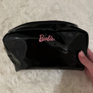 Svart Barbie kosmetiska väska med dragkedja - Cool svart kosmetiska väska från Barbie i glansigt syntetmaterial med rosa Barbie-logga på framsidan. Väskan har en rektangulär form och stängs med en guldfärgad dragkedja upptill. Perfekt för att hålla koll på dina småsaker.