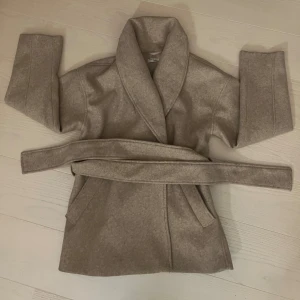 Beige omlott kappa med bälte - Säljer en beige kappa med bred krage och knytskärp i midjan. Jackan har två fickor framtill och är i ett mjukt ull-liknande material. 