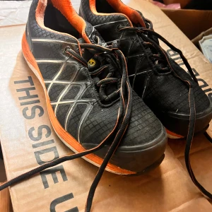 Svarta och orange sneakers från Gesto - Säljer ett par svarta sneakers från Gesto med orange detaljer och innersula. Skorna har snörning, mönstrad sula och coola vita linjer på sidorna. Perfekta för dig som gillar sportig stil och vill sticka ut med färg. Storlek 39.