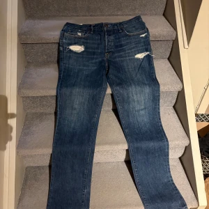 Abercrombie & Fitch slitna jeans - Säljer ett par Abercrombie & Fitch jeans i mörkblå tvätt med slitningar och hål på framsida och baksida. Klassisk femficksmodell med orange sömmar och rak/taper fit. Jeansen har en snygg, lite använd look i 100% bomull. Storlek 34/34!Helt nya! Kom privat för frågor eller bud☺️