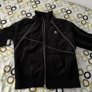 Svart track jacket  - Svart track jacket från Björn Borg med vita detaljer och dragkedja framtill. Jackan har vita ränder på ena ärmen, diskret logga på bröstet och quiltat foder med mönster. Materialet är mjukt och glansigt, troligen polyester. Perfekt för en sportig och clean look.