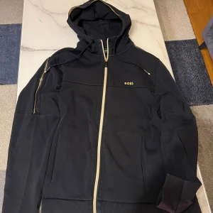 Svart hoodie från BOSS med guldiga detaljer - Svart hoodie från BOSS med dragkedja och huva. Guldiga detaljer längs dragkedjan, huvan och axlarna samt BOSS-logga på bröstet. Tillverkad i mjuk bomull, perfekt för en chill och stilren look.