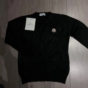 Svart stickad tröja från Moncler - Svart stickad tröja från Moncler med rund halsringning och Moncler-logga broderad på bröstet. Klassisk och stilren modell med ribbade muddar vid ärmslut och nederkant. Perfekt för dig som gillar exklusiva och tidlösa plagg.