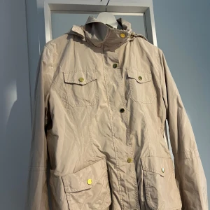 Beige parkajacka från Massimo Dutti - Snygg beige parkajacka från Massimo Dutti med huva, fyra fickor med guldiga knappar och dragkedja framtill. Jackan har en loose passform och är gjord i lätt syntetmaterial, perfekt för vår och höst. Stilren design med coola detaljer.