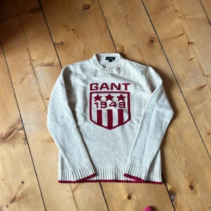 Beige stickad tröja från GANT - Beige stickad tröja från GANT med röd logga och sköld framtill. Tröjan har ribbade muddar med röda detaljer vid ärmslut och nederkant. Klassisk rund halsringning och långärmad modell, perfekt för kyligare dagar.