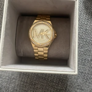 Äkta Guldig Michael Kors  - Säljer en snygg guldig Michael Kors klocka med bred länk och stort MK-logo på urtavlan. Urtavlan är dekorerad med glittrande stenar runt kanten och har en lyxig känsla. Kommer i original ask med Michael Kors logga. Perfekt för dig som gillar bling och statement-accessoarer.
