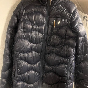Svart dunjacka Peak Performance S - Snygg svart dunjacka från Peak Performance i storlek S. Använd 1-2 gånger. Jackan har vågigt quiltad design, huva och praktiska fickor med dragkedja. Perfekt för kalla dagar och har en sportig look med diskret logga på bröstet.