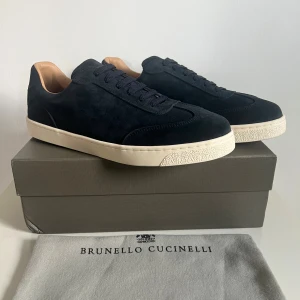 Mörkblå sneakers från Brunello Cucinelli - Snygga mörkblå sneakers från Brunello Cucinelli i lyxig mocka med vita platta sulor och mörkblå snörning. Skorna har en stilren och minimalistisk design med rund tå och diskret branding på plösen. Perfekta för dig som gillar exklusiva och tidlösa sneakers.