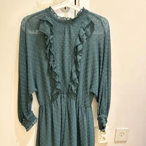 Ny  kortklänning med volanger - Supersöt blå kortklänning med långärmade ärmar och volangdetaljer längs bröstet och ärmsluten. Klänningen har ett diskret prickigt mönster och är gjord i ett lätt, transparent tyg som ger en luftig känsla. Perfekt för dig som gillar romantisk och trendig stil.