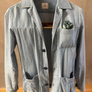 Ljusblå jeansjacka från John Henric - Snygg ljusblå jeansjacka från John Henric med klassisk krage, tre stora fickor och knäppning framtill. Jackan har lång ärm och en avslappnad passform. Perfekt för dig som gillar stilren och enkel design.