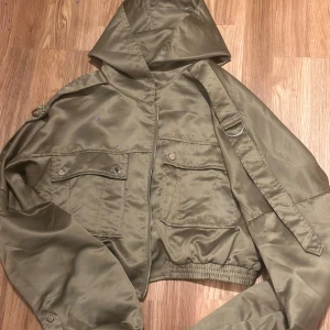 Olivgrön croppad jacka PrettyLittleThing - Snygg olivgrön croppad jacka från PrettyLittleThing med huva, dragkedja och stora fickor framtill. Jackan har coola detaljer som axelremmar med metallring och elastisk midja. Perfekt för dig som vill ha en streetstyle-vibe.