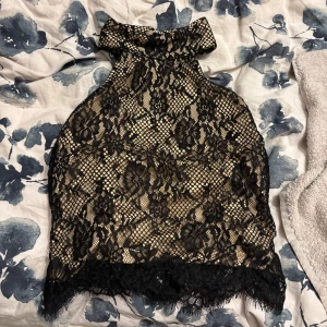 Svart spetsig halterneck topp - Säljer en snygg halterneck topp med svart spets och nude-färgad underdel. Toppen har hög krage, knäppning i nacken och vackra blommiga spetsdetaljer. Perfekt för dig som vill sticka ut med en elegant och trendig look.