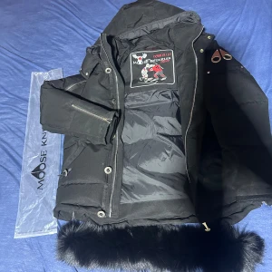 Svart dunjacka Moose Knuckles - Säljer en svart dunjacka från Moose Knuckles med avtagbar pälskant på huvan och snygga metalldetaljer på ärmen. Jackan har dragkedja, knappar och innerfoder med broderad patch. Perfekt för kalla dagar och riktigt stilren look.