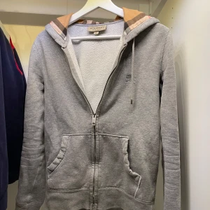Grå Burberry hoodie XS - Grå hoodie med dragkedja från Burberry i bommul, den klassiska logga broderad på bröstet och det ikoniska rutmönstet i luvan. Tröjan har två fickor framtill, snörning i huvan och långa ärmar. Perfekt för en chill och stilren look. Mvh Hannes!