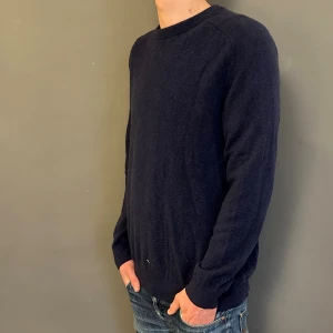 Mörkblå stickad tröja i kashmir - Mysig mörkblå stickad tröja i kashmir med rund halsringning och långa ärmar. Enkel och stilren design som passar perfekt till jeans eller chinos. Tröjan har en normal passform och är supermjuk mot huden.