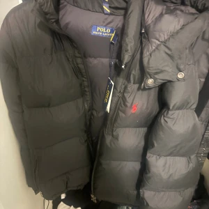 Svart pufferjacka från Polo Ralph Lauren - Svart pufferjacka från Polo Ralph Lauren med huva och dragkedja. Jackan har diskret röd logga broderad på bröstet och är vadderad för extra värme. Klassisk och stilren design med stora paneler och praktiska fickor. Kontakta vid frågor 