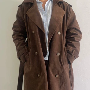 Brun fauxmocka trenchcoat med bälte - Snygg brun trenchcoat i fauxmocka med dubbelknäppning, bred krage och bälte i midjan. Jackan har långa ärmar, axelklaffar och klassiska detaljer som ger en cool och tidlös look. Perfekt för dig som vill sticka ut med en stilren och trendig jacka.