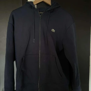 Mörkblå hoodie från Lacoste - Snygg mörkblå hoodie från Lacoste med dragkedja och klassisk krokodil-logga på bröstet. Perfekt för en avslappnad stil. Den har långa ärmar och en praktisk huva med dragsko.