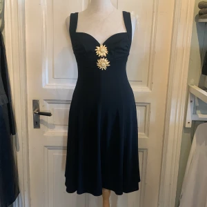 Svart klänning med guldblommor - Supersnygg svart klänning med breda axelband och markerad byst. Klänningen har två stora dekorativa guldblommor framtill som ger en unik och cool touch. Modellen är utsvängd och går till knäna, perfekt för dig som vill sticka ut med en elegant men lekfull look. Stretchmaterial