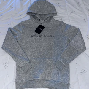 Grå hoodie från Björn Borg - Snygg grå hoodie från Björn Borg med stilrent tryck på bröstet. Tröjan har luva med snörning, ribbade muddar och en stor magficka. Perfekt för chill dagar och enkel att matcha med jeans eller joggers.