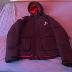 Mörk blå vinterjacka från Henri Lloyd M - Säljer en mörk blå vinterjacka från Henri Lloyd i storlek M. Jackan har röd insida, huva och stora fickor framtill. Perfekt för kalla dagar med sin rejäla och sportiga look. Klassisk logga på bröstet och detaljer på ärmen.