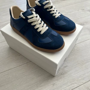 Maison Margiela blå  - Snygga sneakers från Maison Margiela i marinblå mocka och skinn med vita snören och beige innersida. Skorna har en klassisk gummisula i brunt och stilren design som passar till det mesta. Perfekta för dig som gillar exklusiva och trendiga sneakers. Det är bara att skriva om du har frågor!
